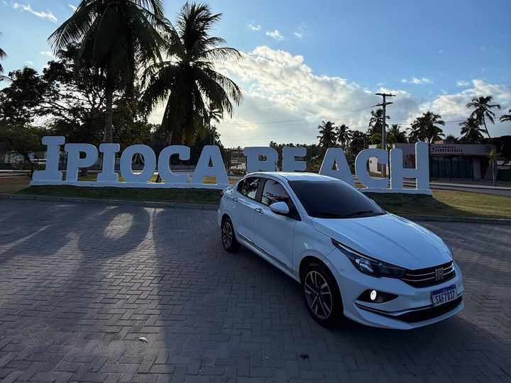 Carro branco do translado estacionado em frente ao letreiro Ipioca Beach, em Maceió